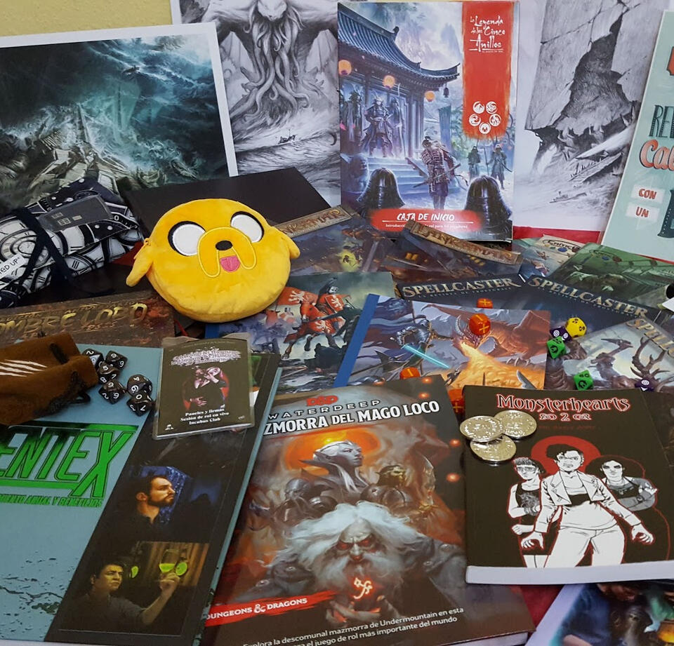 Fotografía de una mesa llena con varios de los libros en los que he trabajado y algún adorno, como un monedero con Jake o mi pase para el evento de Vampiro: la Mascarada.